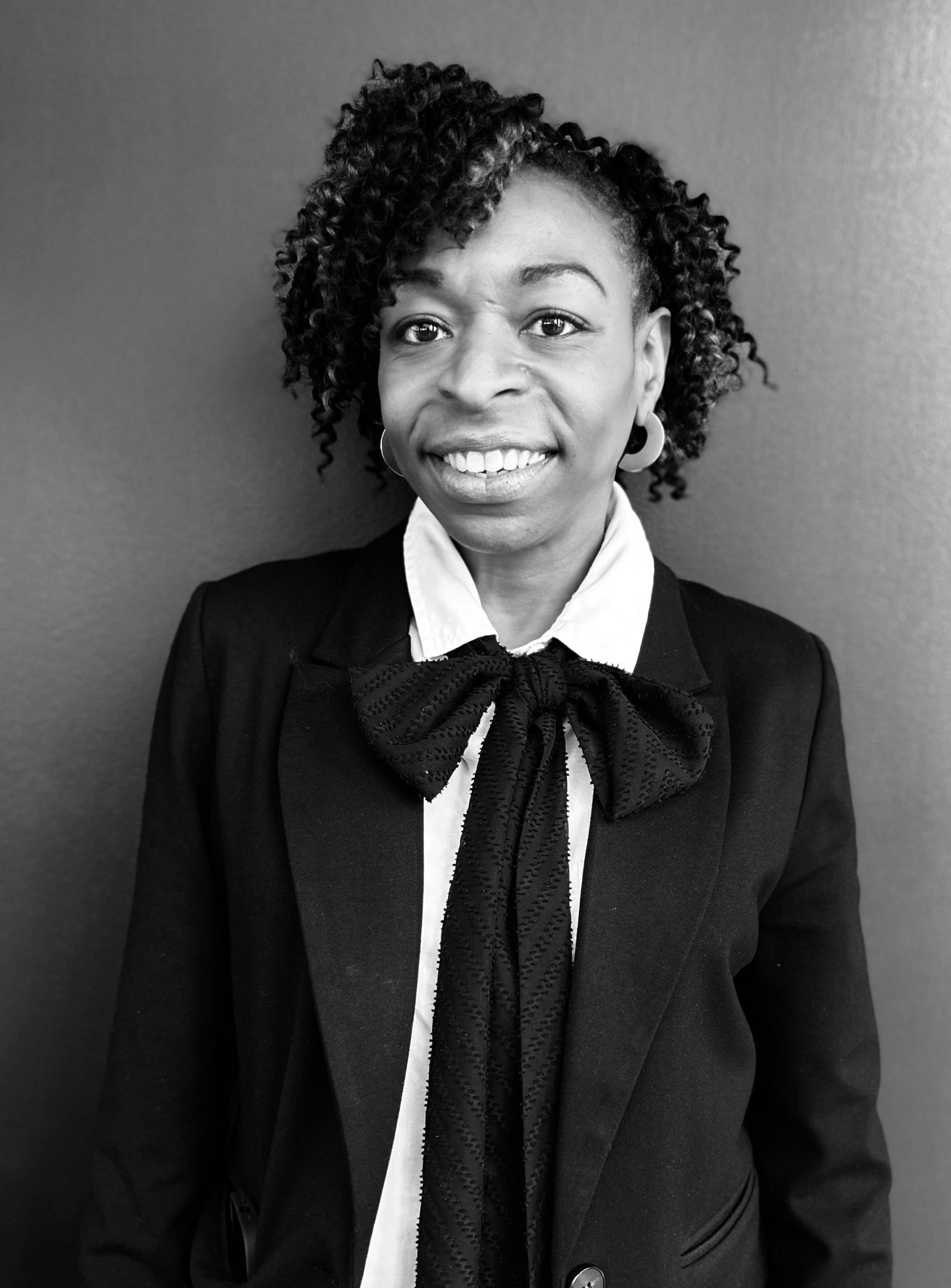 Portrait of a smiling professional in a black suit and bow tie against a plain background, exuding confidence.